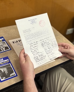 Hands holding a typed letter with handwritten notes and a red stamp, viewed above a table with pamphlets and materials related to Edwin Pittman.