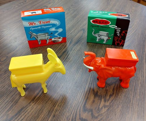 The box on the right shows an illustration of the elephant cigarette dispenser with a cigarette being dispensed out the rear of the animal and a man smoking a cigarette. The caption of the box reads: Mr. G.O.P. Cigarette Dispenser. What do you think of the campaign promises? Pull my trunk and see!! The plastic elephant is red in color with white tusks.
The box on the left shows an illustration of the donkey cigarette dispenser with a cigarette being dispensed out the rear of the animal and a cigarette sitting in an ash tray. The caption of the box reads: Mr. Dem. Cigarette Dispenser. What do you think of the campaign promises? Pull my ears and see!! The plastic donkey is entirely yellow in color. 